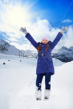 Happy 8 years old Caucasian girl in blue standing with lifted from excitement hands on snow outside in winter