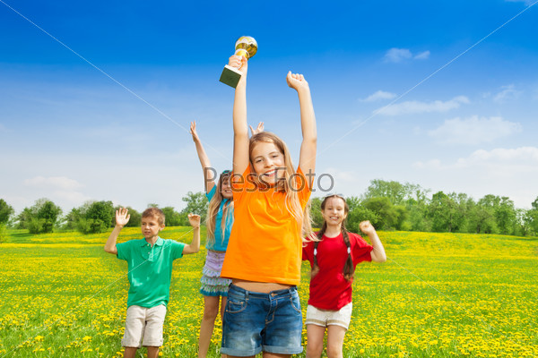 Portrait of happy little girl holding prize in lifted hands cup with her team cheering on background