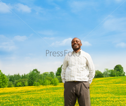 Happy confident laughing black man wearing shirt standing outside in the park on the yellow dandelion field