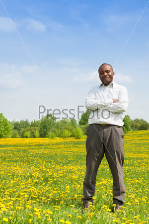 Happy confident black man wearing shirt standing outside in the park on the yellow dandelion field