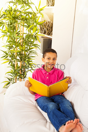 Nice black 5 years old boy sitting on the couch with yellow book and reading