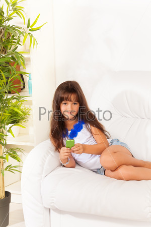 Happy shy smiling little girl with cell phone text her friends laying on the white leather couch