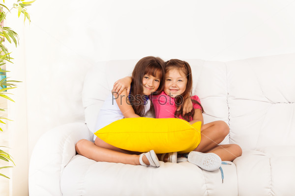 Two happy, Asian and Caucasian calm and relaxed girls years old sitting and hugging holding pillow on the white leather coach in living room at home
