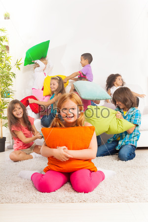 Happy smiling little girl hugging pillow with large group of her friends fighting with pillow on the coach
