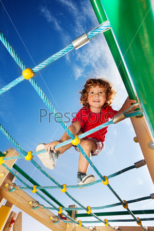 Happy little smiling three years old child boy climbing on the playground ropes with sky on background