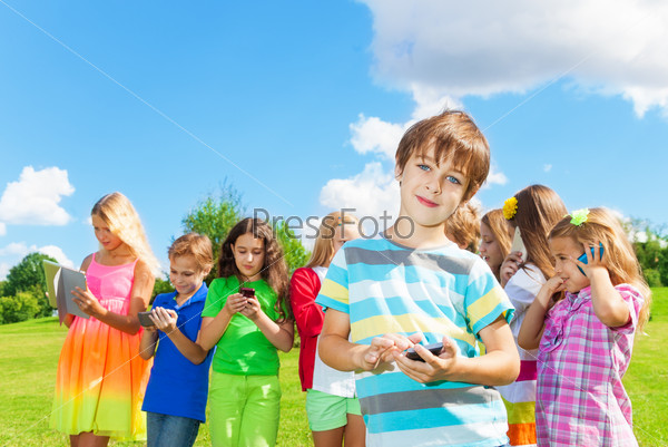 One smiling school age boy standing with digital devices, text, sms, play, post to social network outside, with group of happy friends kids boys and girls