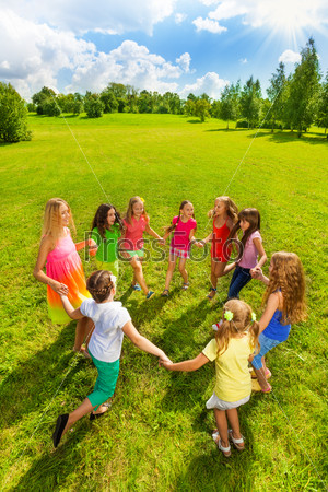 Large group of happy girls play roundelay and stand in circle in the park on the green grass on sunny summer day top view