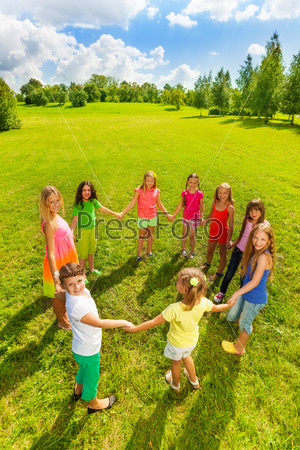 Large group of happy girls play roundelay and stand in circle in the park on the green grass on sunny summer day, view from above