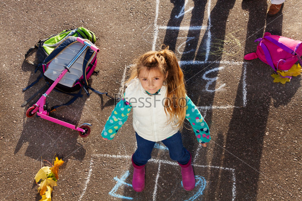 Nice little girl jumping over hopscotch game after school with bags and scooter laying near