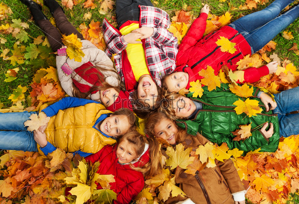 Large group of kids laying in the grass with maple leaves all over them on autumn day