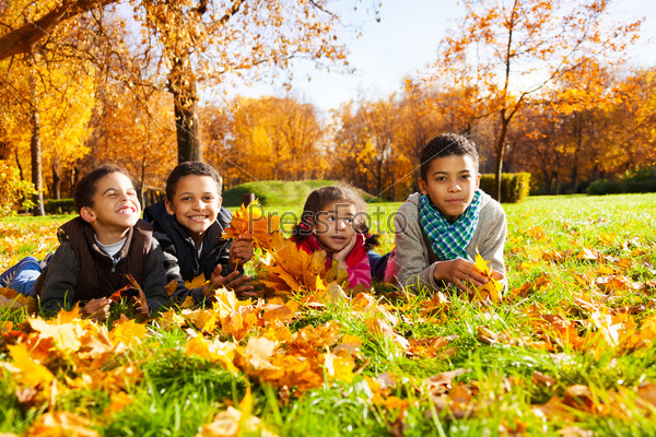 Group of four boys and girl, happy brothers and sister 3-10 years old laying in grass together  in the park in autumn clothes holding maple leaves bouquets