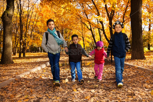 Group of four boys and girl, happy brothers and sister 3-10 years old going together holding hands in the park wearing backpacks and autumn clothes in oak park with orange leaves
