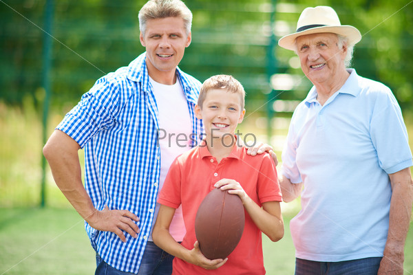 Cute boy with rugby ball and his grandfather and father looking at camera outdoors