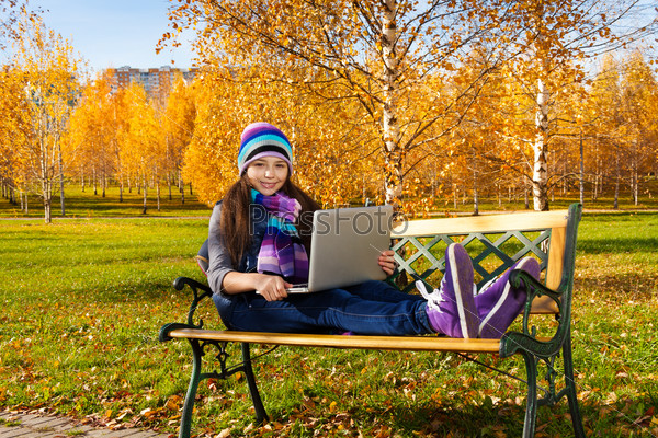 Beautiful school girl sitting on the bench in park with laptop top on sunny autumn day
