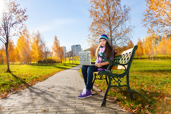 Beautiful school girl 11 years old sitting on the bench in park with laptop top on sunny autumn day wearing warm clothes, stock photo