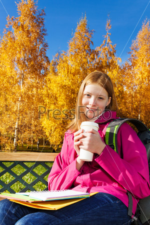 Beautiful teen 14 years old school girl sitting on the bench in autumn park holding coffee and textbooks