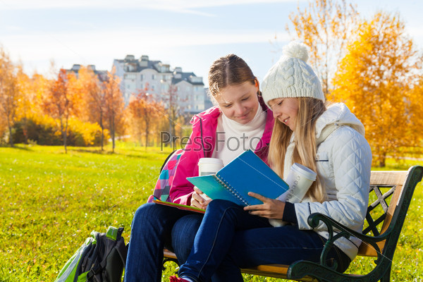 Two happy 14 years old girls in the autumn park taking about homework holding textbook and coffee