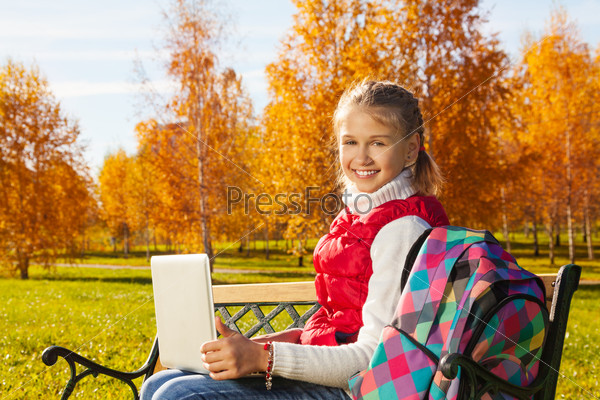 Happy blond 11 years old girl with amazing smile sitting on the bench with laptop turning back doing homework outside in the autumn park on sunny day