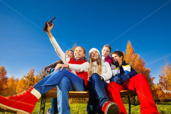 Wide angle shoot of group of five children sitting on the bench and self photographing with camera phone, boy and girls on autumn sunny day