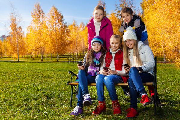 Five happy school age kids sitting on the bench with mobile cell phones happy and smiling, boy and girls on sunny autumn day wearing warm clothes