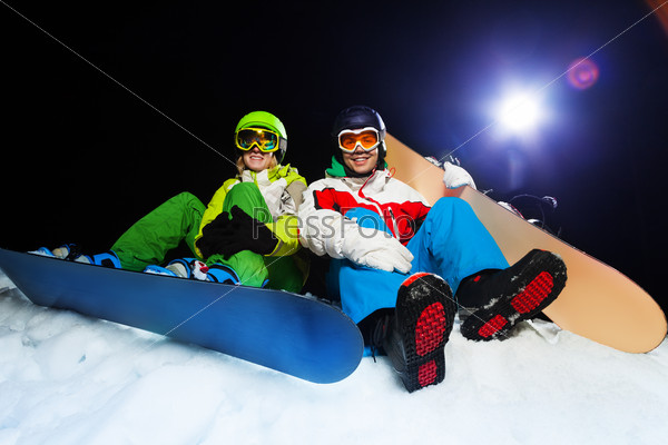 Two smiling snowboarders wearing ski masks sitting together\
at night with flash on the background