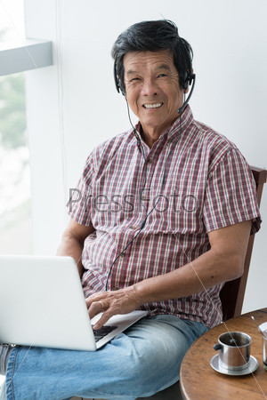 Portrait of cheerful senior man in headset working on laptop