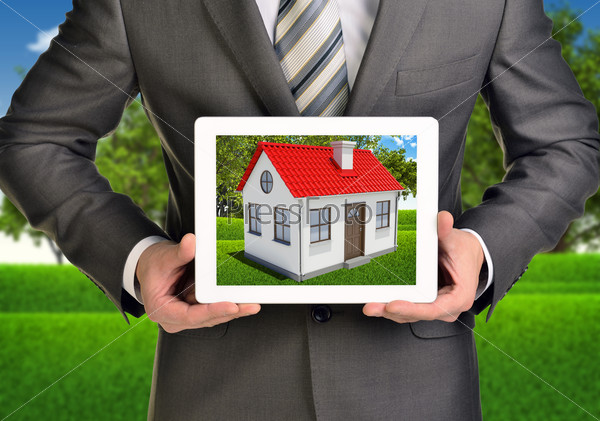 Hands hold tablet pc. Picture of small house with red roof on screen. Blurred landscape as backdrop, stock photo
