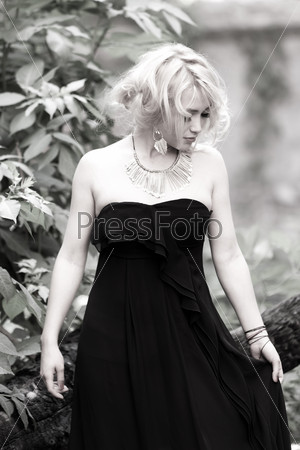 A black and white image with a beautiful and delicate blonde woman in black long dress, looking down while holding one side of it.
