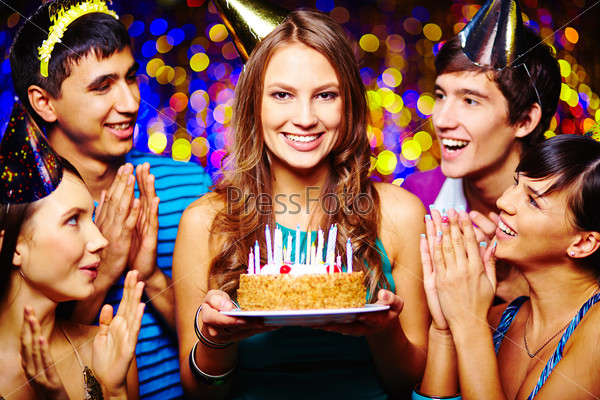 Beautiful young female with birthday cake looking at camera, her friends applauding