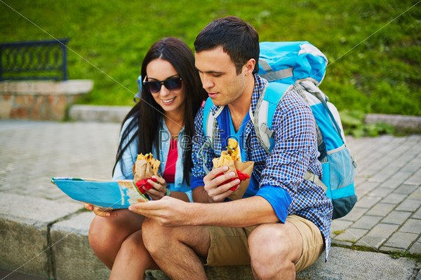 Young travelers looking at map while sitting on pavement and eating fast food