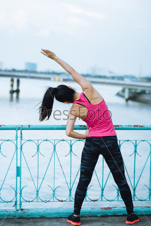 Rear view of female jogger doing side bends standing against a bridge railing