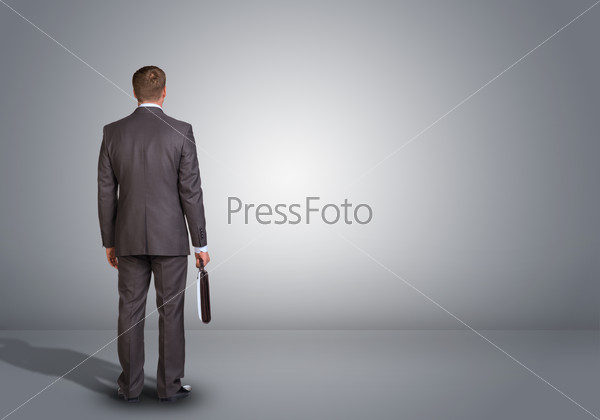 Businessman in suit standing in an empty gray room. Rear view. In hand holding briefcase