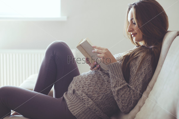 Home cozy portrait of pregnant woman resting at home and reading book on sofa, stock photo