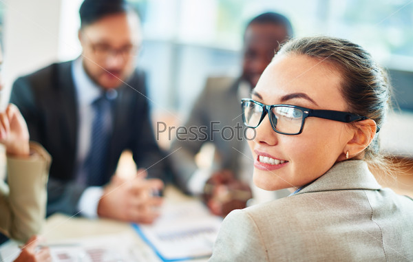Portrait of beautiful secretary in eyeglasses looking at camera on background of co-workers