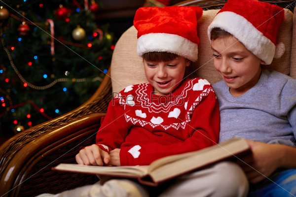 Adorable siblings in Santa caps reading book together on Christmas evening