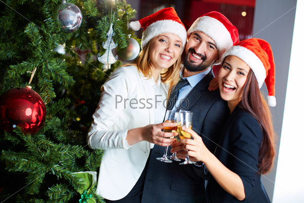 Friendly co-workers in Santa caps toasting with champagne by Christmas tree in office
