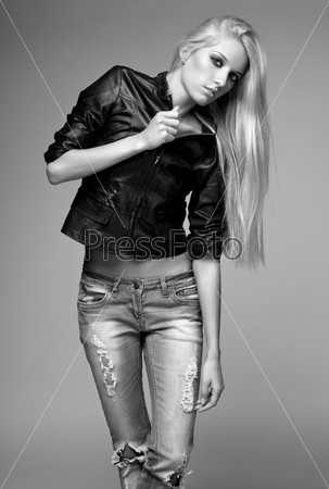 Monochrome portrait of blonde young woman in ragged jeans and black jacket on gray background