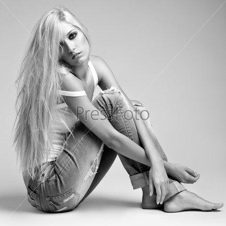 Monochrome portrait of blonde young woman in ragged jeans and vest sitting on floor on gray background