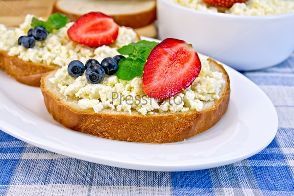 Slices of bread with curd cream, mint, blueberries and strawberries, a bowl of cottage cheese, bread on a board on a background of the blue checkered tablecloth