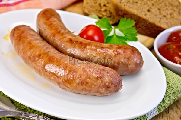 Pork sausages fried in a dish, bread, sauce, tomato, parsley, fork, knife, napkin on the background of wooden boards
