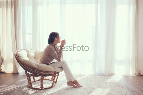Young woman at home sitting on modern chair in front of window relaxing in her lliving room and drinking coffee or tea, stock photo