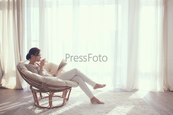 Young woman at home sitting on modern chair in front of window relaxing in her lliving room reading book and drinking coffee or tea