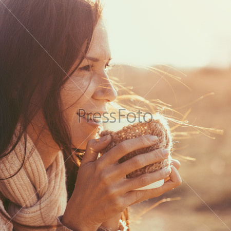 Woman wearing warm knit clothes drinking cup of hot tea or coffee outdoors in sunlight, instagram toned, square composition