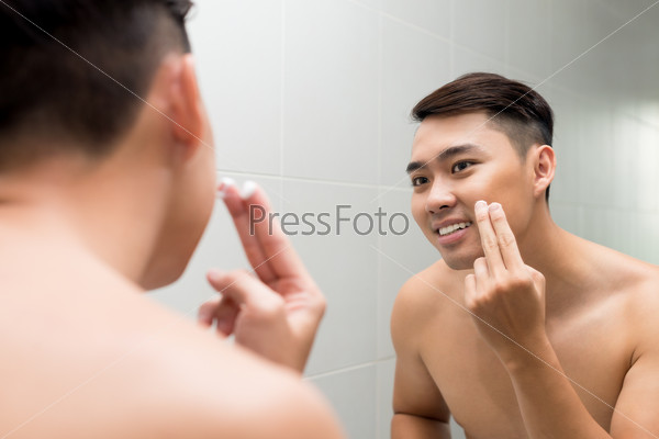 Young man applying facial lotion in front of the mirror