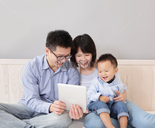 A happy, family of mother, father, son sitting on bed at home having fun using a tablet computer pc, asian people