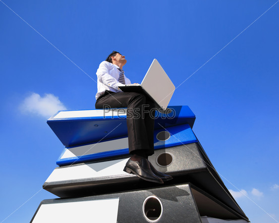 Happy business man using notebook computer and sitting on lots piles of document folders, technology concept