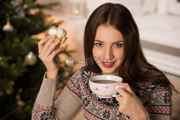 Beautiful woman wearing winter outfit drinking tea with candy at home near Christmas tree