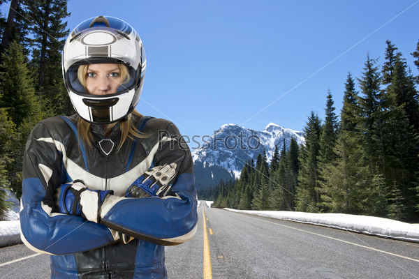 biker chick, wearing a motorcyclist suit and helmet, posing on a snowy road in beautiful mountain scenic, surrounded by large pine trees and the melting snow in the warm spring sun
