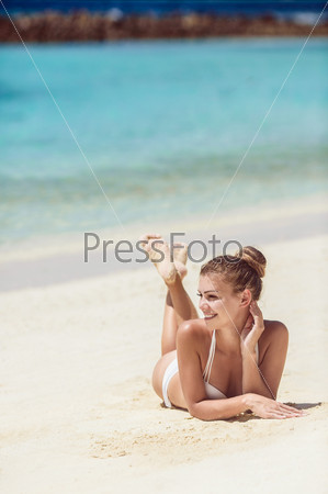 An attractive young woman wearing a white bikini laying on a beach with her elbow on the sand. Ocean can be seen in the background. Vertical shot.