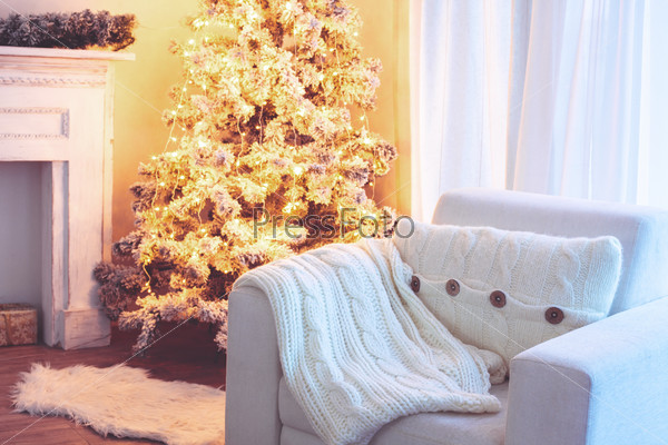 Beautiful holiday decorated room with Christmas tree and white comfortable chair with soft knitted blanket and cushion on it